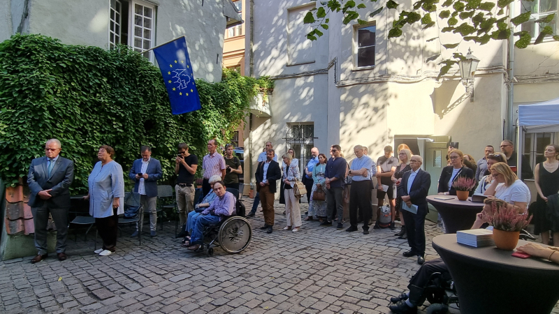 Bruģēts iekšpagalms ar vēsturisku noskaņu. Vīteņiem apaudzis mūris, Eiropas kultūras mantojuma dienu karogs, sīkrūtiņu logi, vasarīgs laiks. Pie stāvgaldiņiem un apkārt atrodas dažādi vasarīgi ģērbušies vīrieši un sievietes, arī vairāki cilvēki ratiņkrēslos.