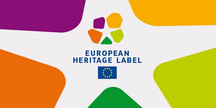 Oficiālais Eiropas mantojuma zīmes logo - taisnstūris, tā sānos ir oranžas, lillā, zaļas un sarkanas krāsas figūras, centrā atkārotas tās pašas krāsas neregulāras formas daudzstūros, zem tām angļu valodā, drukātiem burtiem zilā krāsā rakstīts European heritahe lable, apakšā ir Eiropas savienības karogs. Fons ir balts.