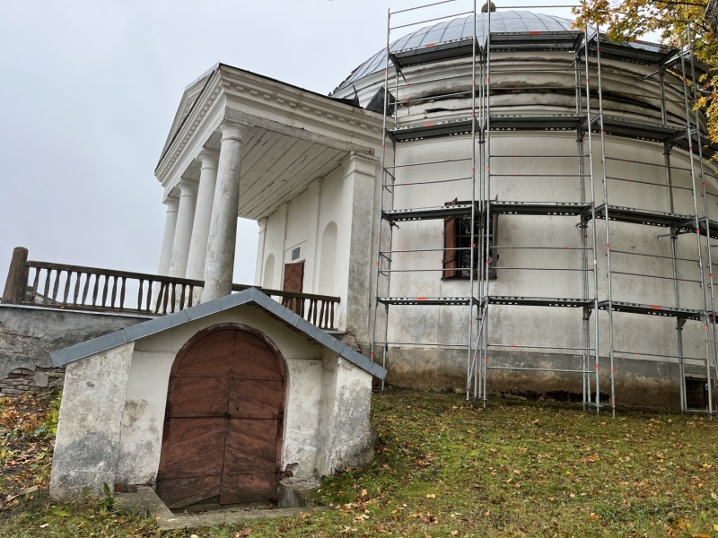 Fotogrāfija, kurā redzama Varakļānu muižas kapliča jeb Svētā Viktora kapela, kas atrodas restaurācijas procesā. Būves apaļā daļa pa labi ir apjozta ar metāla sastatnēm. Kapela ir gaišā krāsā ar tumšu kupola jumtu. Priekšplānā pa kreisi redzama mazāka piebūve ar brūnām koka durvīm un nobružātām sienām. Ap kapelu zeme klāta ar zaļu zāli un rudenīgām lapām, un apkārtnes koki ir ar retām lapām, kas norāda uz vēsāku laiku. Debesis ir apmākušās.