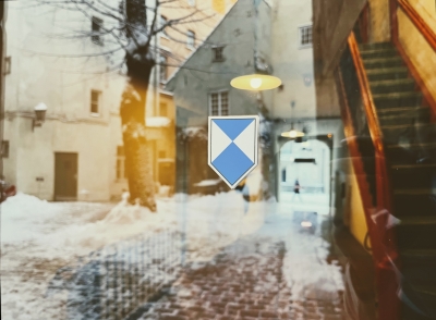 Attēla centrā ir zilā vairoga zīme - zili balts vairoga formas apzīmējumus, ko izvieto pie kultūras pieminekļiem. Zīme ir uzlīme, kas pielikta pie caurspīdīgām stikla durvīm. Foto veikts no ārpuses un stikla durvīs atspoguļojas sniegots pagalms, kura centrā ir koks, bet fonā vēsturiska, balta māja. Caur durvīm ēkā var redzēt apgaismotas, stāvas koka kāpnes. Kāpnes atrodas attēla labajā pusē. 