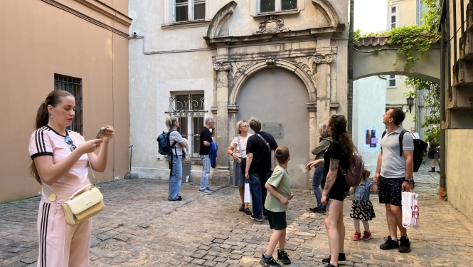 Bruģētā, vēsturiskā pagalmā redzami cilvēki, kas lūkojas visapkārt. Gaišs, vasarīgs vakars.