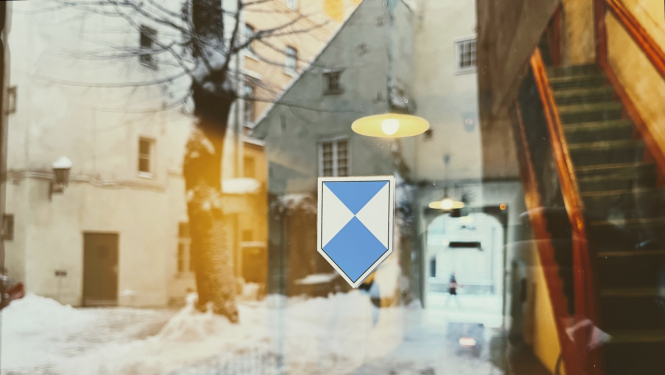 Attēla centrā ir zilā vairoga zīme - zili balts vairoga formas apzīmējumus, ko izvieto pie kultūras pieminekļiem. Zīme ir uzlīme, kas pielikta pie caurspīdīgām stikla durvīm. Foto veikts no ārpuses un stikla durvīs atspoguļojas sniegots pagalms, kura centrā ir koks, bet fonā vēsturiska, balta māja. Caur durvīm ēkā var redzēt apgaismotas, stāvas koka kāpnes. Kāpnes atrodas attēla labajā pusē. 