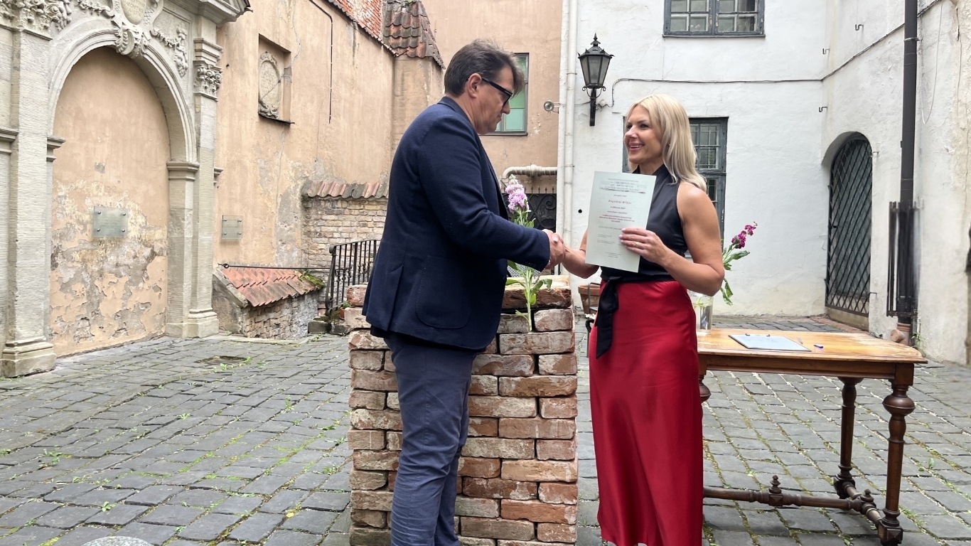 Vīrietis spiež roku sievietei, kura tur rokā diplomu.