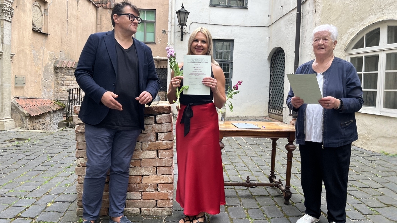 Trīs cilvēki smaidīgi pozē attēlam. Vidū ir sieviete, kas tur diplomu un ziedus.