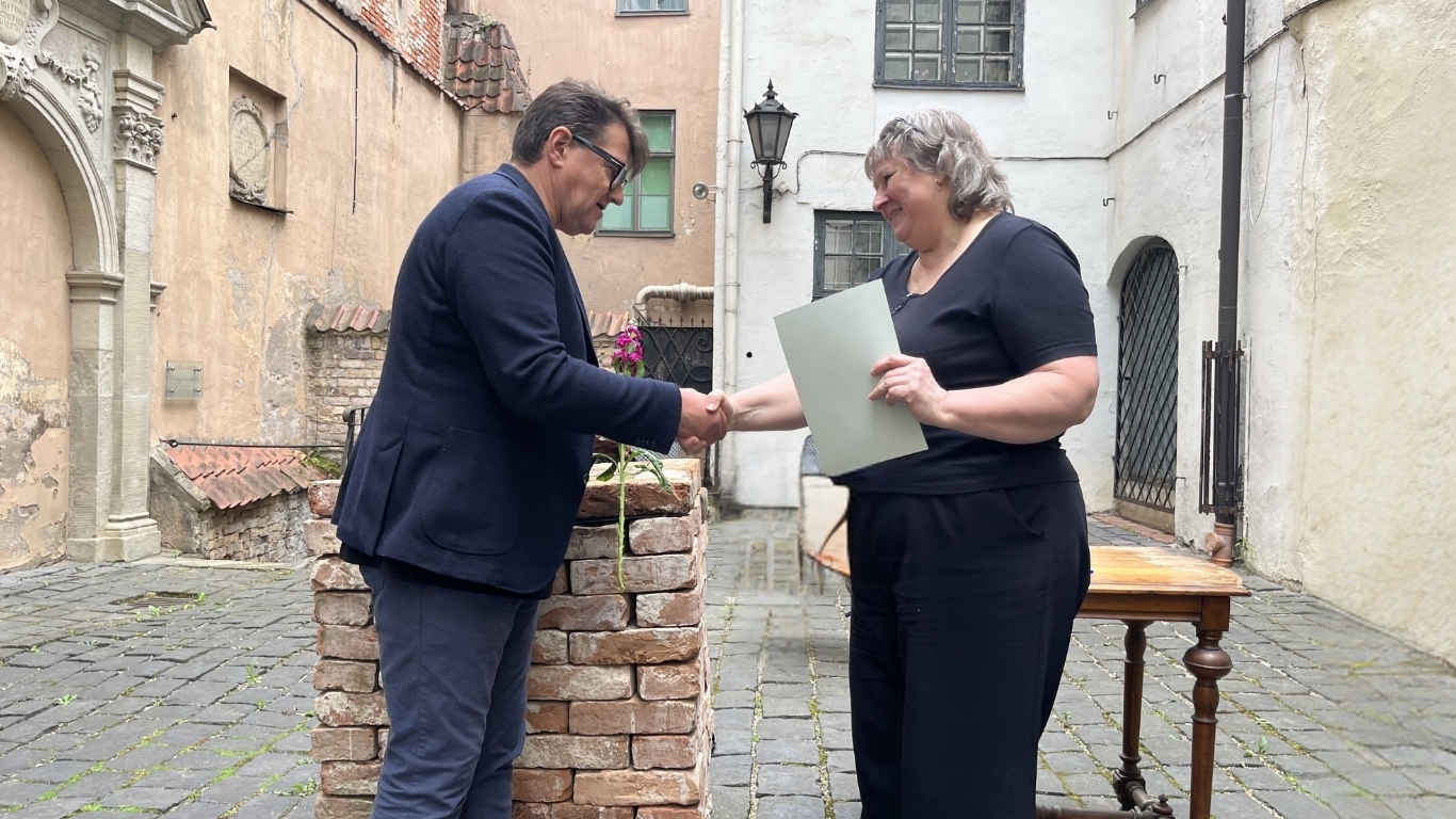 Vīrietis sniedz roku un ziedus sievietei ar diplomu rokā.