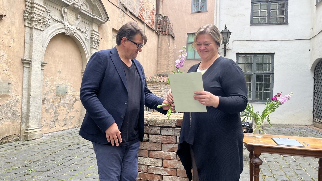 Vīrietis un sieviete stāv svinīgā mirklī, sieviete smaidīgi pēta saņemto diplomu.