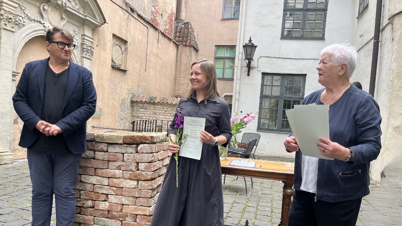Trīs cilvēki svinīgi pozē attēlam. Vidū ir sieviete ar diplomu un ziediem.