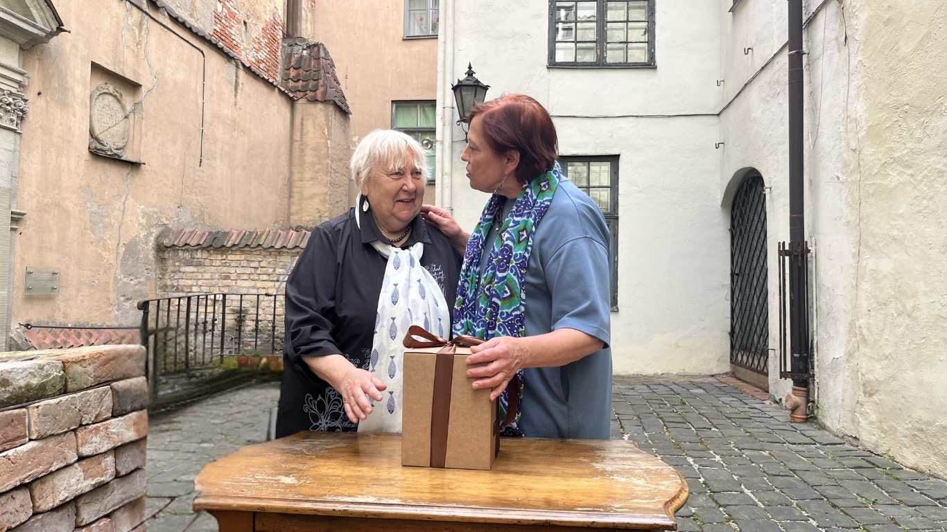 Divas sievietes cieši stāv, viena apsveic otru ar apbalvojumu.