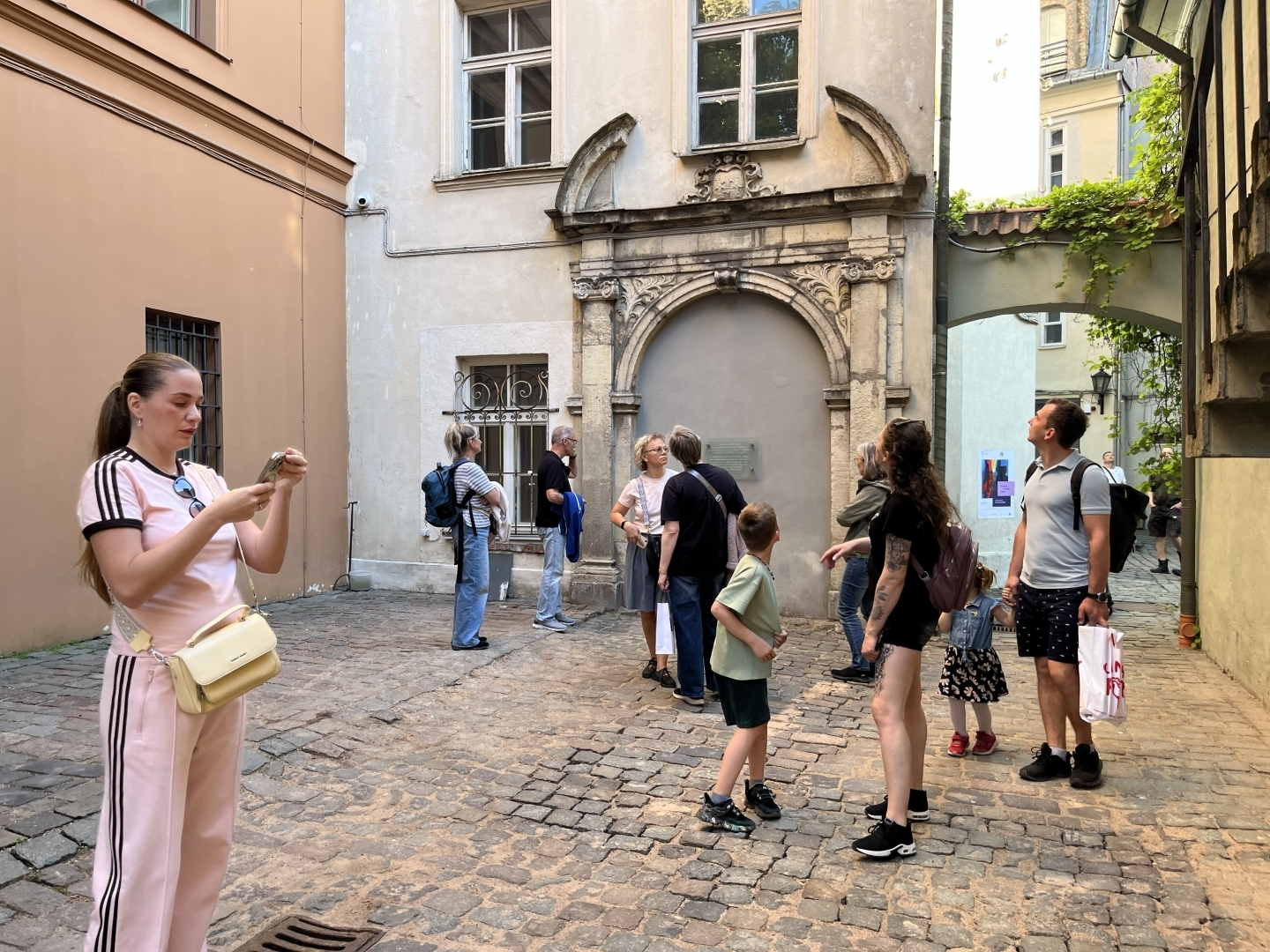 Bruģētā, vēsturiskā pagalmā redzami cilvēki, kas lūkojas visapkārt. Gaišs, vasarīgs vakars.