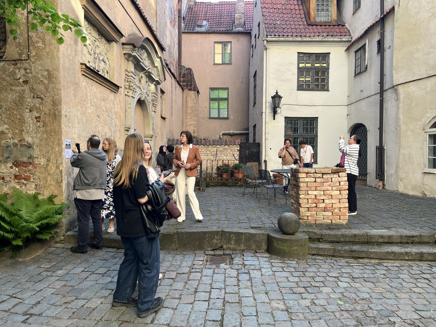 Bruģētā, vēsturiskā pagalmā redzami cilvēki, kas lūkojas visapkārt. Gaišs, vasarīgs vakars.