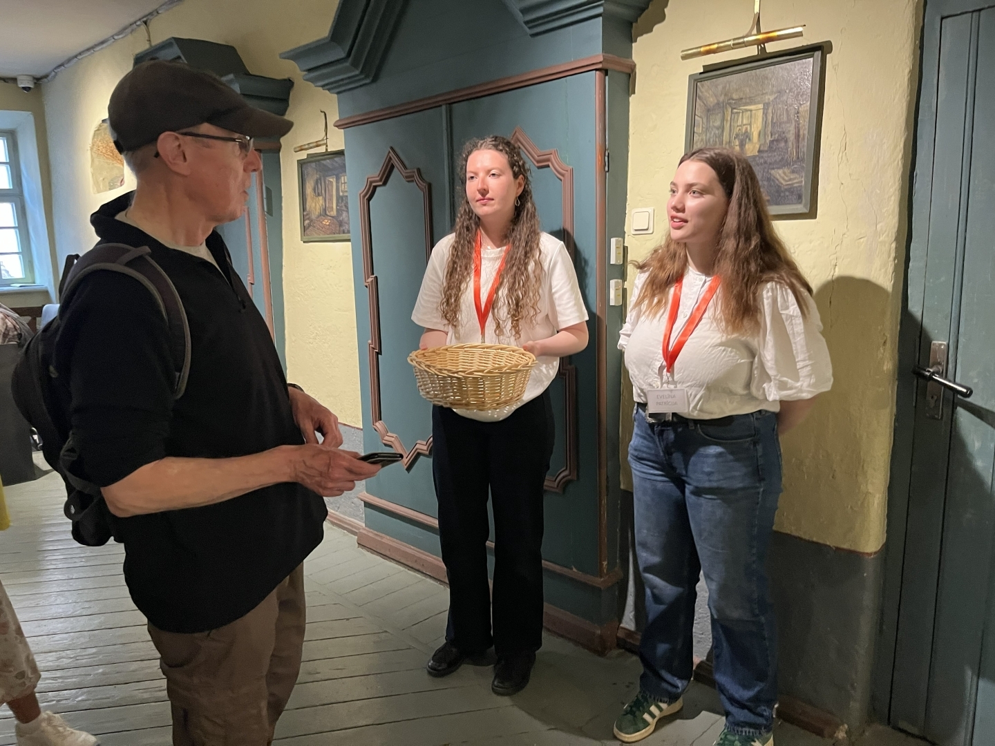 Senatnīgā telpā divas jaunas meitenes ar sarkanās lentās iekarinātām vārdu nozīmītēm runā ar puisi. Vienai no meitenēm rokās ir gaišs, pīts grozs.