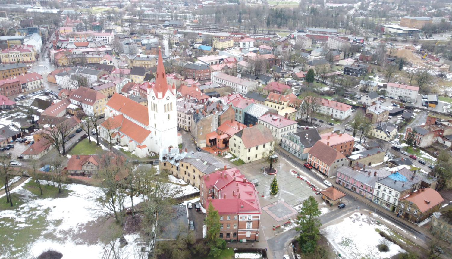 Attēlā redzama Cēsu vecpilsēta no putna lidojuma ziemas periodā. Centrā dominē liela balta baznīca ar sarkanu jumtu un augstu torni, kas ir Cēsu Svētā Jāņa baznīca. Ap to izvietotas vēsturiskas ēkas ar dažādu krāsu fasādēm un sarkaniem vai brūniem jumtiem. Ielas ir šauras un līkumotas, raksturīgas vecpilsētu apbūvei. Priekšplānā redzams laukums ar izrotātu Ziemassvētku eglīti. Dažviet uz zemes un ēku jumtiem ir redzamas nelielas sniega paliekas. Fona daļā redzama mazstāvu dzīvojamā apbūve, koki un dažas ind