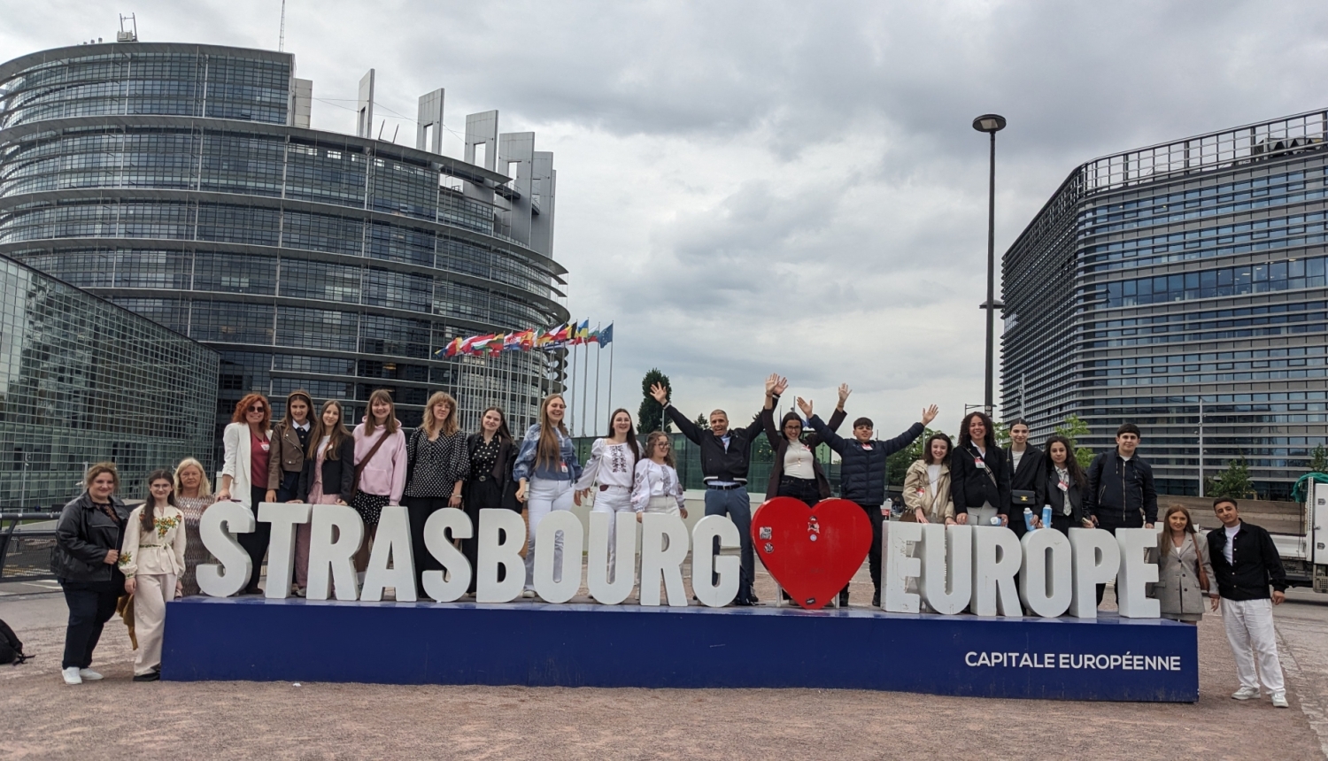 Pilsētvidē izvietots uzraksts STRASBOURG [SIRSNIŅA] EUROPE capitale europeenne. Ap zīmi sastājušies smaidīgi jaunieši un daži pieaugušie, kopā 22 cilvēki, ģērbušies ikdienšķās drēbēs. Fonā Eiropas parlamenta ēka. Debesis ir pelēkas.