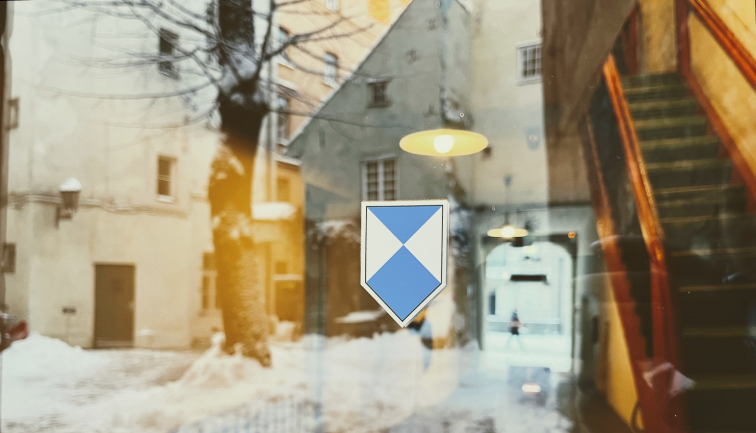 Attēla centrā ir zilā vairoga zīme - zili balts vairoga formas apzīmējumus, ko izvieto pie kultūras pieminekļiem. Zīme ir uzlīme, kas pielikta pie caurspīdīgām stikla durvīm. Foto veikts no ārpuses un stikla durvīs atspoguļojas sniegots pagalms, kura centrā ir koks, bet fonā vēsturiska, balta māja. Caur durvīm ēkā var redzēt apgaismotas, stāvas koka kāpnes. Kāpnes atrodas attēla labajā pusē. 