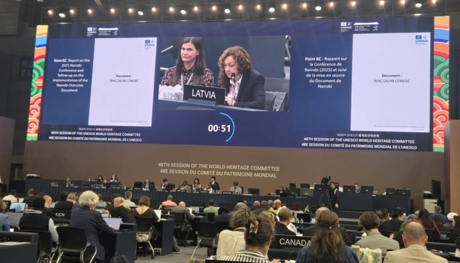 No lielas konferenču telpas aizmugures noķerts skats ar cilvēku mugurām un pašā priekšā lielu ekrānu, kurā redzama Latvijas delegācija - divas sievietes. Skatuve ir tālu no sēdētāja, tāpēc to, ka viņas sēž zāles priekšā var tikai nojaust