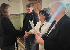 Četras personas stāv pasākuma telpā. Sievietes kreisajā pusē sniedzas rokasspiedienā ar sievieti centrā, kura tur papīrus. Vīrietis melnā uzvalkā un vecāka sieviete stāv blakus, vērojot.