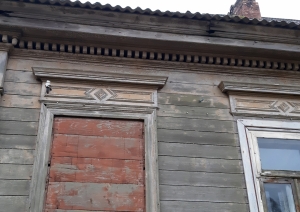 Attēlā redzami divi 19.gs. koka muižas ēkas logi, viens ir aizsists ar dēļiem, otrā ir logs. Fasādē redzami kokgrebumi.
