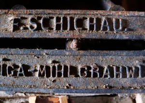 Tuvplāna attēls vecam, rūsas dzelzs mehānisma elementam. Uz detaļas ir reljefs uzraksts: augšējā rindā 'F. SCHICHAU.' un apakšējā rindā 'RIGA-MÜHLGRABEN' (Rīga-Mīlgrāvis). Starp abām uzrakstu rindām atrodas horizontāla sprauga. Detaļa ir netīra un veca, ar tumšu, raupju tekstūru.