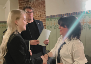 Jauna sieviete (kreisajā pusē) uzvalkā sarokojas ar sievieti (labajā pusē) baltā kreklā un kaklasaitē. Aiz viņām stāv vīrietis melnās drēbēs, turot papīrus.