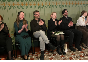 Klātesošie aplaudē sēžot sešos krēslos pasākuma zāles priekšplānā. Sienas ir dekorētas ar zaļiem zīmējumiem. Centrā redzama jauna sieviete, kura tur rokā rozā rozi.