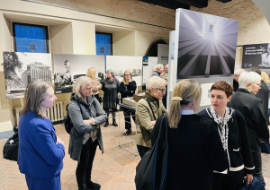 Cilvēki izklaidus pa telpu tumšos un zilos apģērbos sarunājas izstāžu zālē. Fonā pie sienām izvietotas lielformāta melnbaltas arhitektūras fotogrāfijas. Telpai ir redzami vēsturiski logi ar nelielām rūtīm.