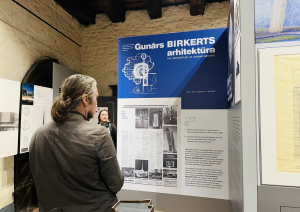 Vīrietis ar gariem, astē sasietiem pelēcīgi brūniem matiem no mugurpuses vēro lielu, zilu informatīvu stendu ar uzrakstu 'Gunārs Birkerts arhitektūra'. Uz stenda redzami ēku rasējumi un teksti.