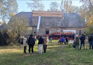 Attēlā redzama Ālaves kungu māja no attāluma, pie mājas notiek pasākums un priekšplānā redzamas skatītāju muguras.