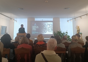 Attēls veikts no telpas aizmugures. Tajā redzamas cilvēku muguras, bet tālumā, zāles priekšā redzams gara auguma runātājs, kurš rāda uz ekrāna redzamu prezentāciju.