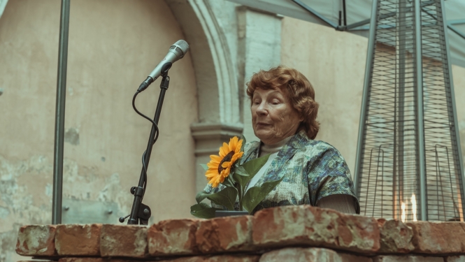 Centrā redzama kundze gados, viļņotiem matiem, svinīgās drēbēs. Priekšplānā no ķieģeļiem sakrauta tribīne, mikrofons, viņa tur rokā saulespuķi. Fonā - gāzes lampa (āra apkurei) un vēsturisks protāls.