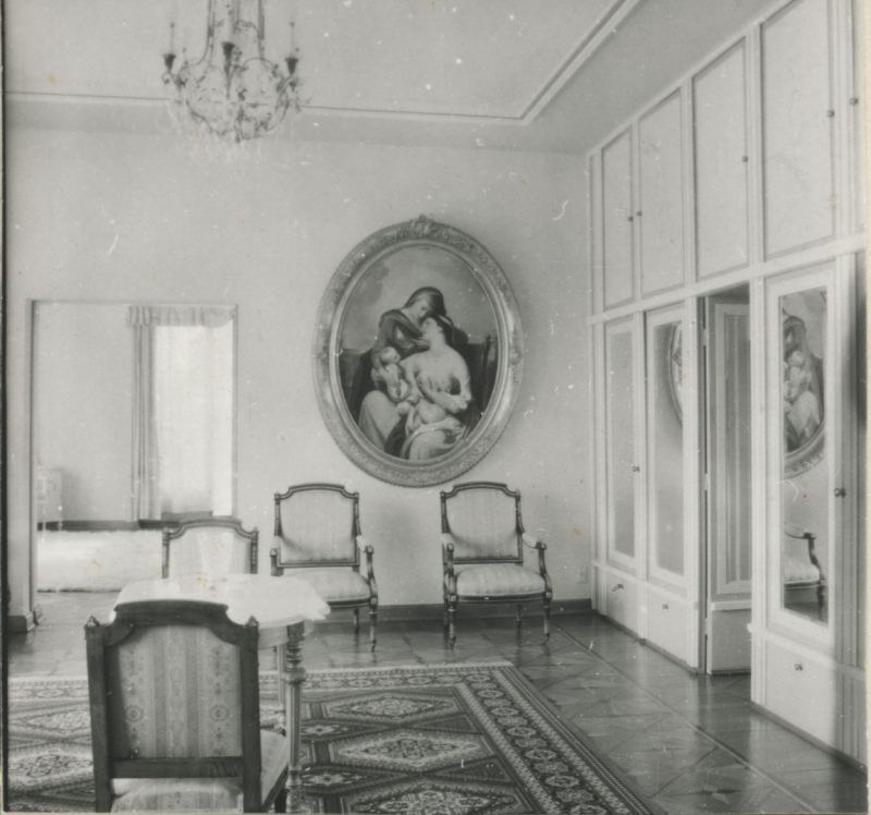 Grezns telpas interjers ar gleznu pie tālākās sienas, četriem krēsliem un sneu galdu, labajā pusē durvis.