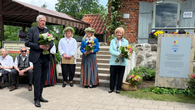 Uz betona bruģa celiņa stāv četri cilvēki - garš, sirms kungs tumšā uzvalkā ar puķu pušķi un trīs sievietes - divām galvā ir puķu vainadziņi, vidējā tērpusies latviešu tautas tērpā, sievietēm rokās ir pušķi. Cilvēki stāv blakus Eiropas mantojuma zīmes plāksnei. Fonā redzamas vēsturiskas ēkas - sarkani ķieģeļi un dakstiņi. 