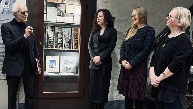 Attēla kreisajā pusē kungs sirmiem matiem, melnā žaketē ar brillēm aktīvi žestikulējot runā, vēršoties pie trim sievietēm. Sievietes stāv rindā attēla labajā pusē, ir svinīgi ģērbušās tumšās drēbēs. Visas trīs rokas tur sev priekšā sakrustotas.  Fonā vēsturiska mūra siena un vitrīna ar izstādes materiālu. Apakšā redzama sarkanu ķieģeļu grīda. Telpa ir tumši apgaismota, izceļot vitrīnu.