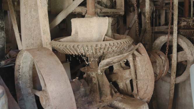 Tuvplāna attēls vecām, putekļainām un rūsas klātām ūdensdzirnavu iekārtām telpā. Centrālajā daļā dominē liels, plats metāla zobrats ar vertikālu asi, kas ir daļa no dzirnavu mehānisma. Abās pusēs redzami lieli rati. Mehānismu klāj putekļi un zirnekļu tīkli, redzams, ka tas ilgstoši nav lietots. Griesti un koka konstrukcijas ap detaļām ir klāti ar biezu gaišu kārtu. Tumšajā fonā redzamas citas iekārtu daļas un ķēdes, kas norāda uz dzirnavu telpas sarežģītību.