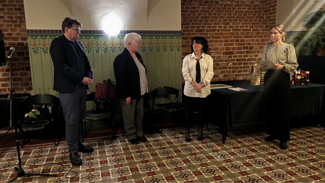 Četras personas stāv zālē, pie neliela galda. Pie galda stāv jauna sieviete un uz galda ir izkārtotas rozes un diplomi. 