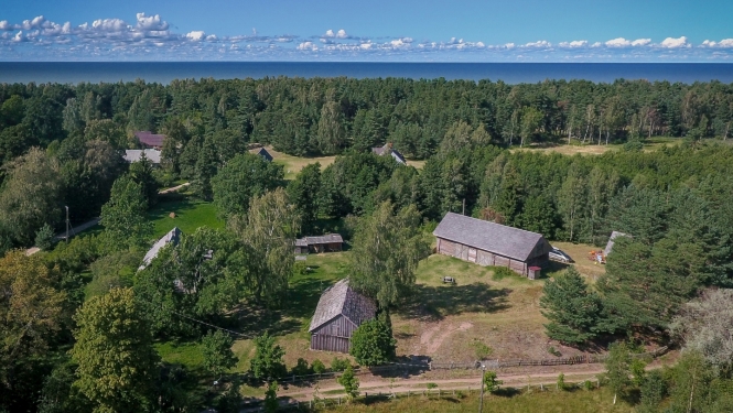 Attēlā redzams pilsētbūvniecības piemineklis - Košraga zvejniekciems. Pilsētbūvniecības piemineklis iekļauj plašāku cilvēka veidotu vidi, kā vecpilsētu vai ciemu. Attēlā redzams ar dronu no gaisa fotografētais Košraga ciems lībiešu krastā. Foto augšpusē pirmo ceturtdaļu aizņem tumši zilas vasaras debesis ar rotaļīgu baltu mākonīšu rindu virs horizonta, kam seko tumši zaļais priežu mežs. Priekšplānā var izšķirt kādu no ciema vēsturiskajām ēkām, starp kokiem šur tur manāmi zemi divslīpju jumti.