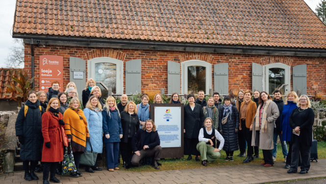 Aptveuni 30 cilvēku grupa pozē fotogrāfijai pie vēsturiskas koka ēkas ar dakstiņu jumtu. Grupa sastājusies ap lielu zīmi, kas ir Eiropas mantojuma zīme. Priekšā ir betona bruģis. Laiks ir rudenīgs.