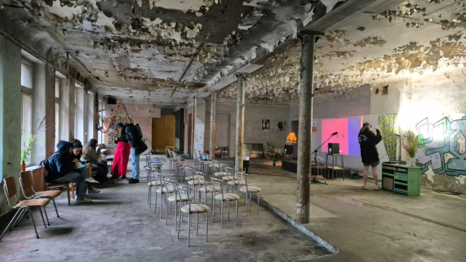 Attēlā redzami studenti. Tie atrodas plašā, neapdzīvotā vēsturiskā telpā. Tai no sienām un griestiem lobās krāsa, izskatās, ka tā tiek izmantota pagaidu pasākumiem. Grīda ir betona. Telpas vidū ir 15-20 metāla krēsli, kas vērsti ar muguru pret logiem. Telpas dziļumā ir neliela skatuve ar 2 mikrofoniem un projektoru, kas projicē rojicē gaiši rozā-purpursarkanu attēlu uz baltas sienas. Skatuves labajā pusē ir grafiti uzraksts, un uz grīdas ir novietotas puķu vāzes. 