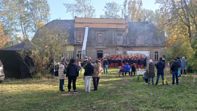Attēlā redzama Ālaves kungu māja no attāluma, pie mājas notiek pasākums un priekšplānā redzamas skatītāju muguras.