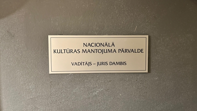 Plāksnīte pie Nacionālās kultūras mantojuma pārvaldes vadītāja durvīm, kas saka "Nacionālā kultūras mantojuma pārvalde"; vadītājs - Juris Dambis. Krāsotās koka durvis ir pelēcīgas, plāksne četrkantīga, neliela, metāla ar melniem burtiem.
