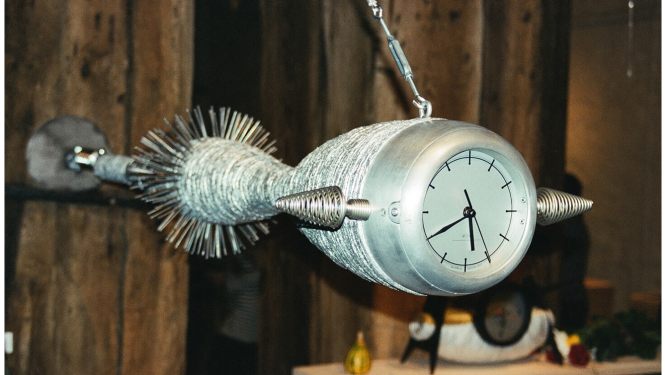 Fotoattēlā redzams dizaina pulkstenis, veidots zemūdenes vai ūdens radības formās ar sudraba krāsas korpusu un baltu ciparnīcu ar melniem rādītājiem. Aiz muguras tam redzama koka dēļu siena. Fotoattēls ir uzņemts no neliela leņķa, un gaisma ir nedaudz izkliedēta.