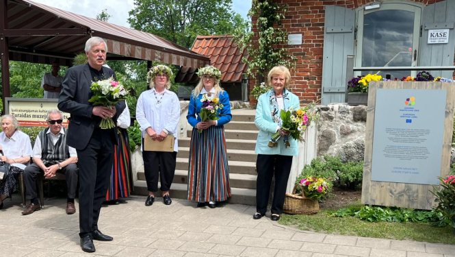 Uz betona bruģa celiņa stāv četri cilvēki - garš, sirms kungs tumšā uzvalkā ar puķu pušķi un trīs sievietes - divām galvā ir puķu vainadziņi, vidējā tērpusies latviešu tautas tērpā, sievietēm rokās ir pušķi. Cilvēki stāv blakus Eiropas mantojuma zīmes plāksnei. Fonā redzamas vēsturiskas ēkas - sarkani ķieģeļi un dakstiņi. 