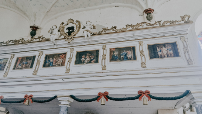 Attēls uzņemts baznīcā,  un tajā redzams balts, grezni dekorēts koka balkons. Balkona augšējā daļā ir rotājumi ar volūtām un figūrām. Centrā ir tumšs, trīsdimensionāls lācis, kas stāv uz četrām kājām. Abpus lācim sēž divas baltas, mazas eņģeļu figūriņas.  Zem augšējā rotājuma redzamas piecas gleznas ar bibliskiem sižetiem, kas ierāmētas zelta rāmjos un izvietotas rindā. Zem gleznām ir platāka koka josla, kuras apakšdaļu rotā tumši zaļa vītne ar diviem sarkaniem, dubultiem bantīšu mezgliem.  Gaismas un ēnas 