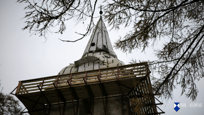 Popes luterāņu baznīcas torņa smaile remontdarbu procesā, fotografēta no apakšas. Tornim noņemts segums, to klāj balts aizsargmateriāls, bet apkārt redzamas koka sastatnes. Smailē redzams krusts un lode, fonā — lapegles zari. Attēls ieturēts tumši pelēcīgos toņos. Labajā apakšējā stūrī pievienota zilā vairoga zīme un baznīcas valsts aizsardzības numurs 6956.