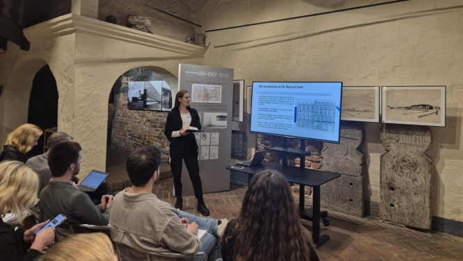 Attēlā redzama vēsturiskas ēkas telpa ar gaišām apmestām sienām, sarkanu ķieģeļu grīdu un brūnām griestu sijām. Kreisajā pusē fonā redzams apvalkdūmenis. Attēla lejasdaļā krēslos redzami cilvēki, kas klausās prezentāciju. Attēla vidū, dziļumā redzama sieviete oficiālā kostīmā, kas kaut ko stāsta un rāda uz digitālo ekrānu, kurā redzama prezentācija.