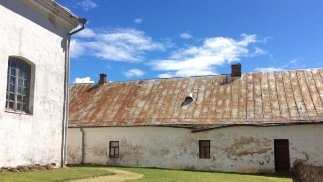 Attēla centrā redzama sena vienstāva ēka ar gaišu fasādi un brūnu jumtu, tai ir nelieli lodziņi. Ēka ir unikālais arhitektūras piemineklis - Rušona klosteris. Sānā pa kreisi, priekšpusē ir balts fasādes fragments, kas ir daļa no blakus esošās baznīcas, pa vidu ir zālājs, attēla augšpusē vasarīgas debesis ar baltiem mākonīšiem.