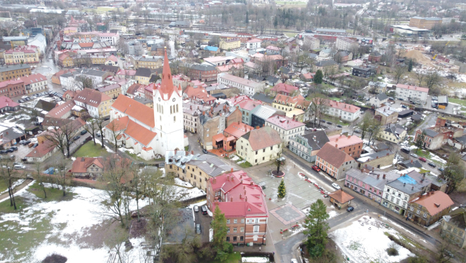Attēlā redzama Cēsu vecpilsēta no putna lidojuma ziemas periodā. Centrā dominē liela balta baznīca ar sarkanu jumtu un augstu torni, kas ir Cēsu Svētā Jāņa baznīca. Ap to izvietotas vēsturiskas ēkas ar dažādu krāsu fasādēm un sarkaniem vai brūniem jumtiem. Ielas ir šauras un līkumotas, raksturīgas vecpilsētu apbūvei. Priekšplānā redzams laukums ar izrotātu Ziemassvētku eglīti. Dažviet uz zemes un ēku jumtiem ir redzamas nelielas sniega paliekas. Fona daļā redzama mazstāvu dzīvojamā apbūve, koki un dažas ind