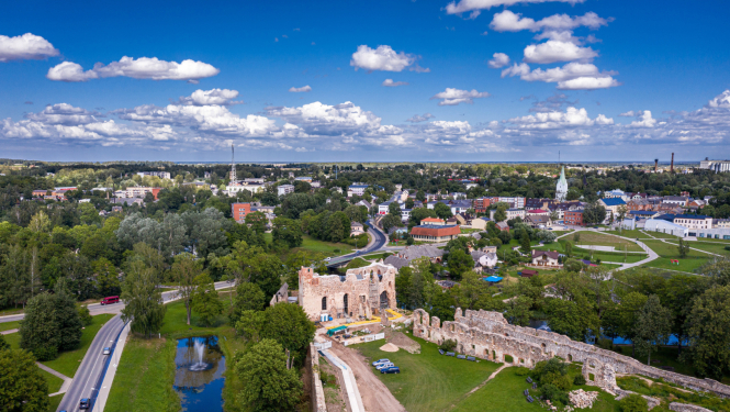 Dobeles pilsētas vēsturiskā centra ainava no putna lidojuma. Centrā priekšplānā redzamas Dobeles pilsdrupas, tām apkārt ir koki un ūdens. Fonā redzama Dobeles pilsētas apbūve, autoceļš un parks. Attēla augšpusē  trešdaļu no attēla aizņem zilas debesis ar baltiem mākoņiem. 