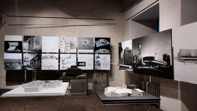 Fotoattēls no izstādes “Birkerts – 20. gadsimta akcenti” iekārtojuma Latvijas Arhitektūras muzejā. Telpā ar vēsturiskām, gaišām mūra sienām un tumšu ķieģeļu grīdu izvietoti arhitekta Gunāra Birkerta darbu eksponāti. Fonā pie sienas divās rindās piestiprinātas kvadrātveida planšetes ar melnbaltām ēku fotogrāfijām, skicēm un rasējumiem. Labajā pusē redzami trīs lielformāta vertikāli paneļi ar ikoniskām arhitektūras fotogrāfijām. Priekšplānā uz metāla karkasa galdiem novietoti divi arhitektūras maketi.