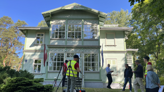 Attēlā redzama vēsturiska, gaiši zaļa koka ēka ar baltiem logu rāmjiem un dekoratīviem kokgriezumiem. Pie fasādes plīvo Latvijas karogs. Ēkas priekšā, zālienā notiek 3D skenēšanas darbi – vairāki cilvēki, daži no tiem dzeltenās atstarojošās vestēs, strādā ar mērniecības aprīkojumu. Kāds vīrietis gaišā kreklā aktīvi skaidro procesu pārējiem, kuri viņu vēro un sarunājas. Apkārt aug zaļi koki, un saulainais laiks rada patīkamu, vasarīgu atmosfēru.
