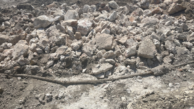 Būvlaukums ar izrakumiem un gružiem – priekšplānā redzami dažāda izmēra akmeņi un betona gabali, zem tiem daļēji atsegta gara, tumša caurule vai koka stienis. Fons ir klāts ar sausu, sablietētu zemi un tehnikas atstātām pēdām.